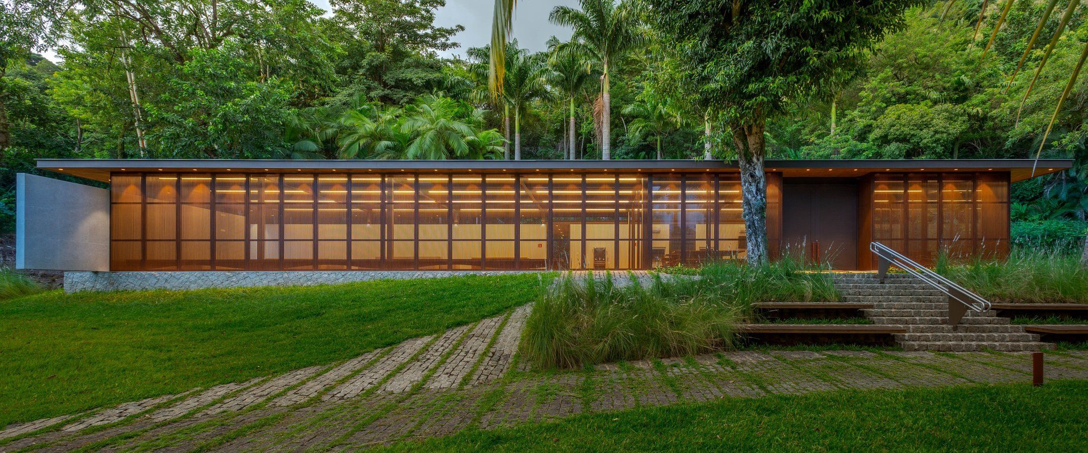 Pavilhão ao entardecer com fachada de brises de madeira iluminada por dentro, projeto Dall'Ovo Magalhães