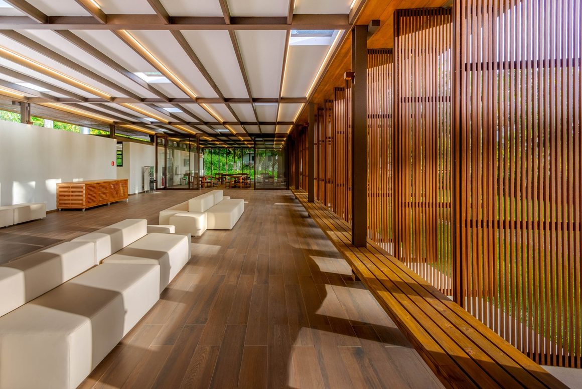 Espaço de convivência com bancos brancos, piso de madeira e painéis de ripas abertos para o jardim