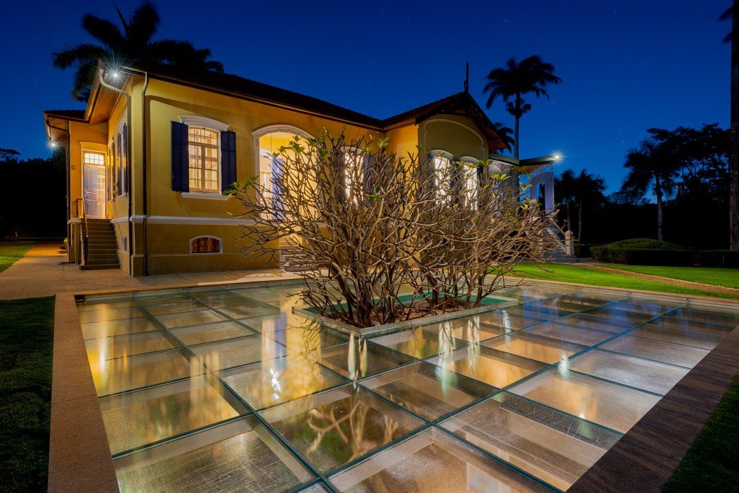 Casarão iluminado à noite, com venezianas azuis e piso de vidro iluminado em volta da árvore